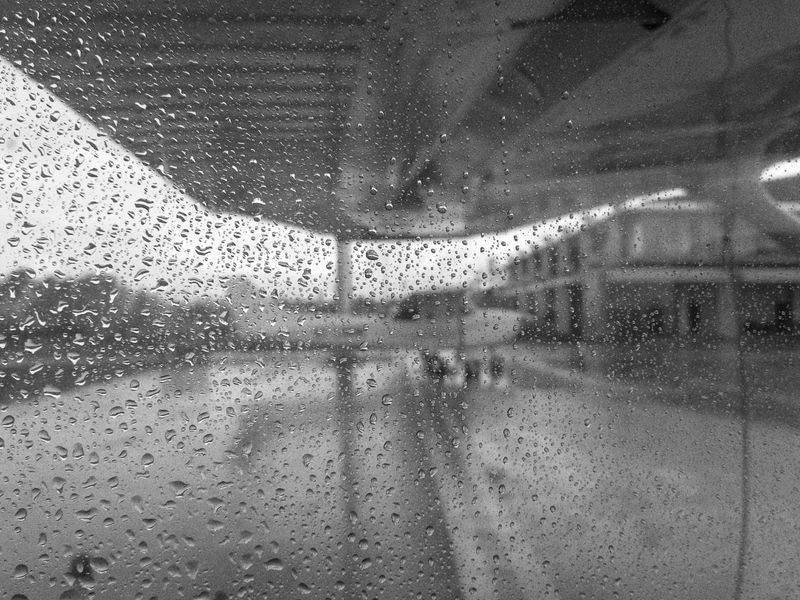 Black and white photograph of raindrops on a window or glass surface, with a blurred airport terminal or hangar visible in the background. Through the rain-covered glass, the silhouette of a small airplane can be seen on the tarmac. The water droplets create a textured foreground that obscures the clear view, giving the image a moody, atmospheric quality.