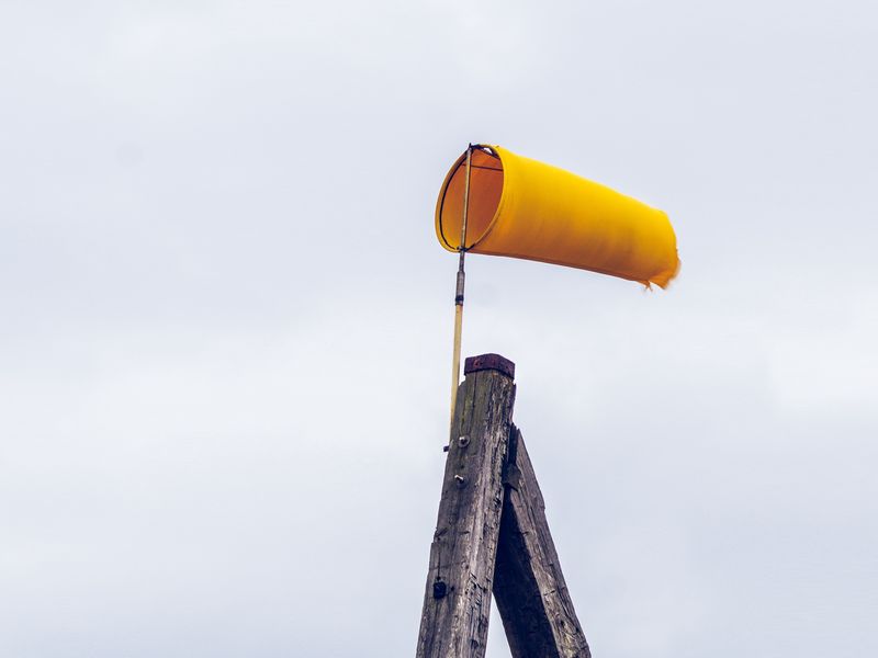 Black and white photograph of raindrops on a window or glass surface, with a blurred airport terminal or hangar visible in the background. Through the rain-covered glass, the silhouette of a small airplane can be seen on the tarmac. The water droplets create a textured foreground that obscures the clear view, giving the image a moody, atmospheric quality.