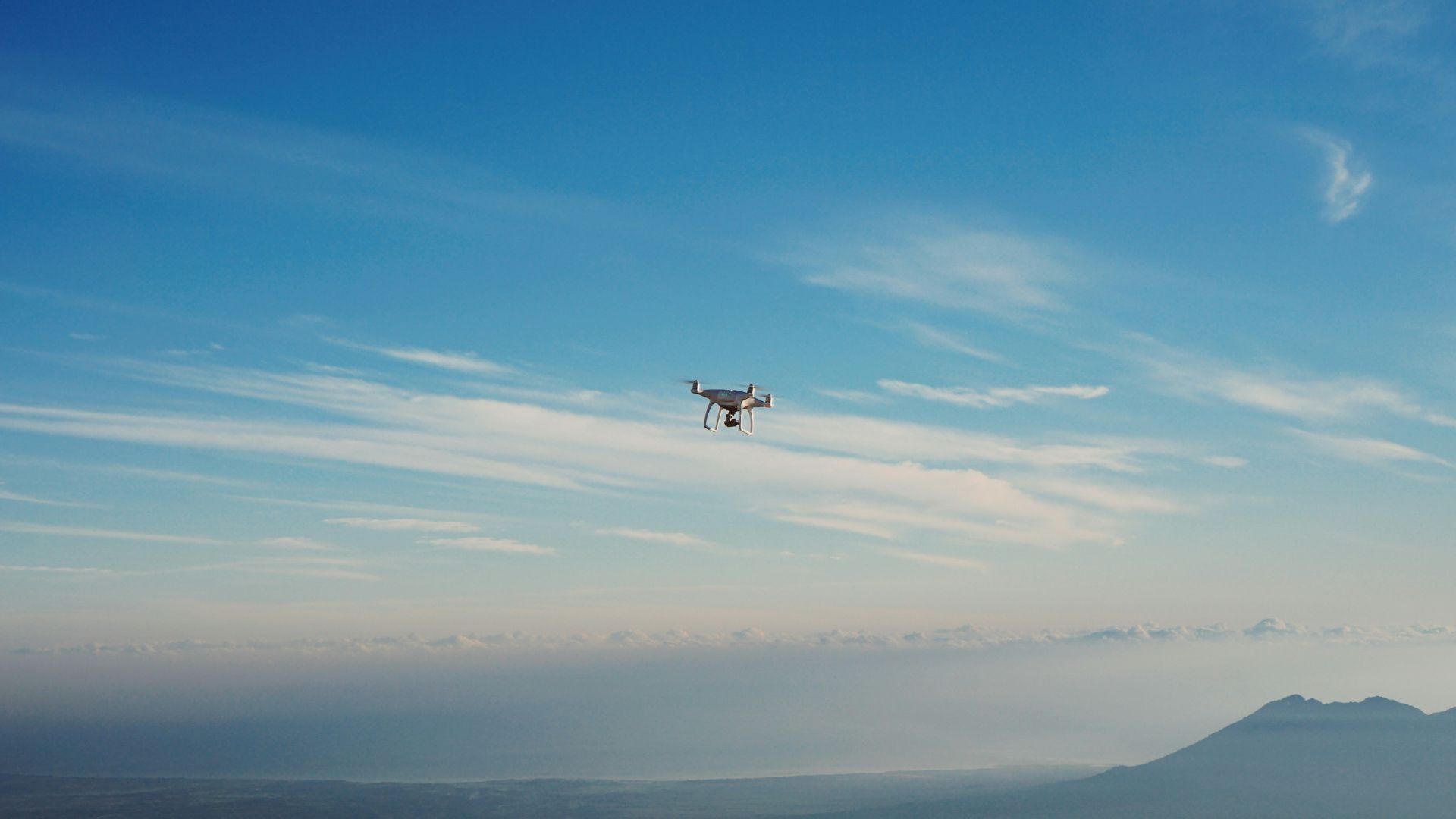 A drone pilot flying a professional camera drone over a scenic landscape with varied terrain. The drone is in flight against a partly cloudy sky, while the pilot monitors the controls with a smartphone/tablet. Beautiful natural scenery stretches below including mountains and water.