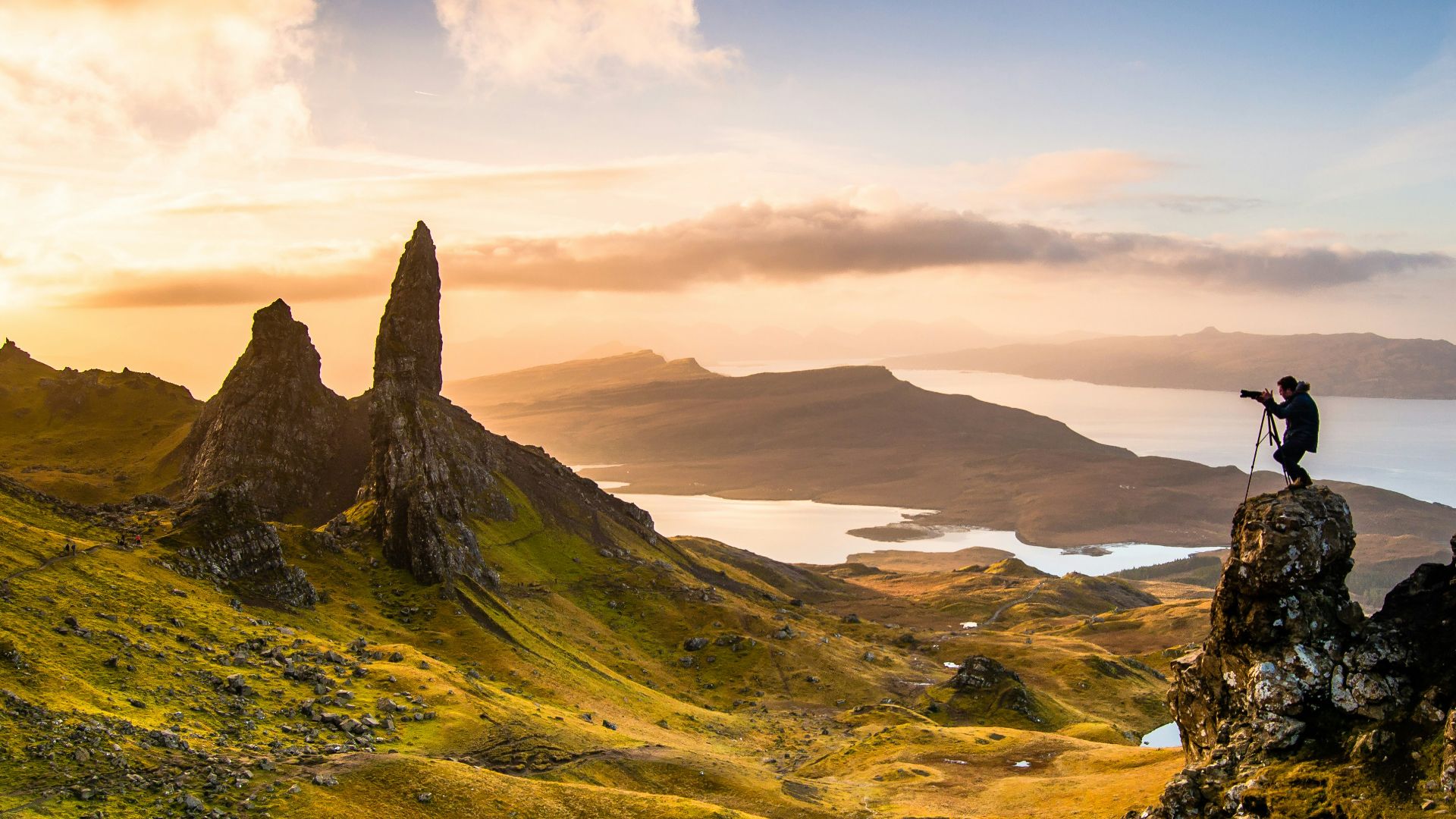 Scenic landscape view at the Isle of Skye in Scotland, showing the distinctive rock formations known as the Old Man of Storr. In golden sunset light, two large rocky pinnacles rise from green hillsides overlooking a distant coastline and mountains. A silhouette of a photographer with a tripod stands on a rocky outcrop capturing the scene.