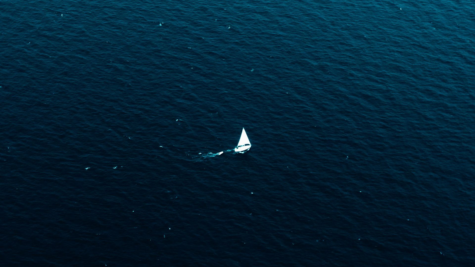 A small white sailboat alone on a vast deep blue ocean, seen from an aerial perspective.