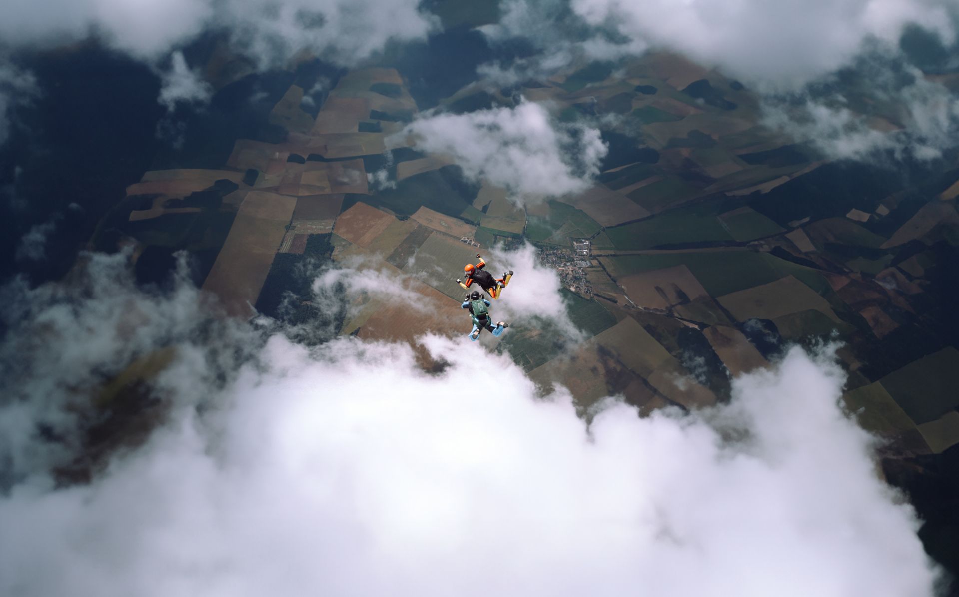 Two skydivers in freefall above farmland with scattered clouds. One skydiver wears a bright orange helmet and black jumpsuit, while the other wears a light blue wingsuit and black helmet. They're performing aerial maneuvers against a backdrop of agricultural fields and patches of clouds.
