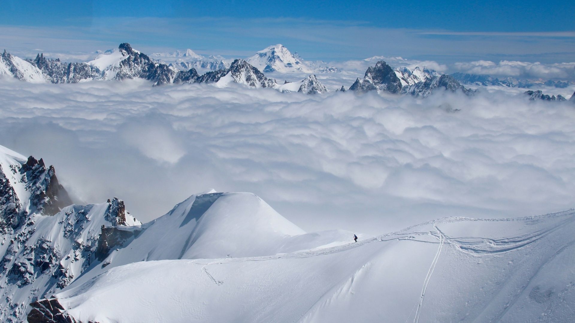 Breathtaking mountain panorama showing snow-covered peaks rising above a sea of clouds, with visible ski tracks and a tiny skier in the foreground on an untouched powder slope.