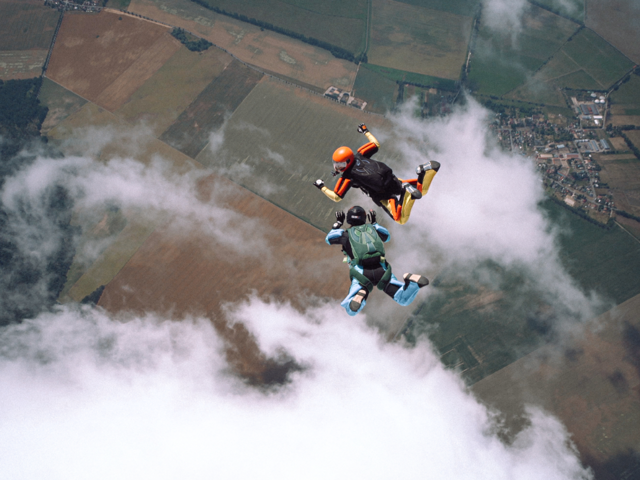 Two skydivers in freefall above farmland with scattered clouds. One skydiver wears a bright orange helmet and black jumpsuit, while the other wears a light blue wingsuit and black helmet. They're performing aerial maneuvers against a backdrop of agricultural fields and patches of clouds.